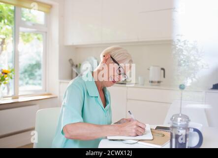 Ältere Frau schreibt im Tagebuch am Morgen Küchentisch Stockfoto