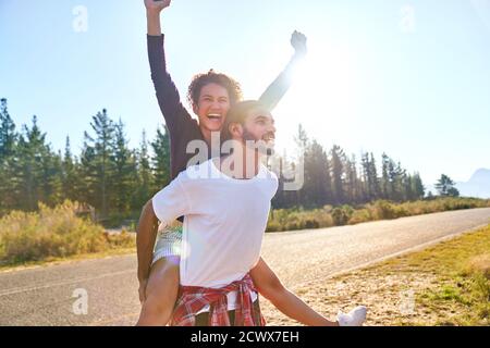 Portrait sorgloses junges Paar Huckepack am sonnigen abgelegenen Straßenrand Stockfoto