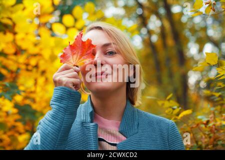 Lächelnde blonde Frau, die ein Auge mit rotem Ahornblatt bedeckt. Herbstkonzept. Im Freien Herbst weibliche Porträt Nahaufnahme Stockfoto