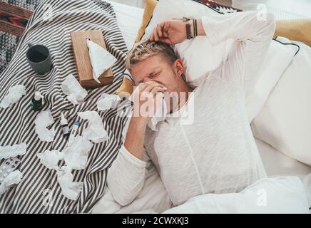 Ungesunder und müder Mann niesen oder wischende notty Nase Mann liegt in einem gemütlichen Bett neben einer Menge von gebrauchten Papiertaschentücher und Medikamente auf der Decke. S Stockfoto