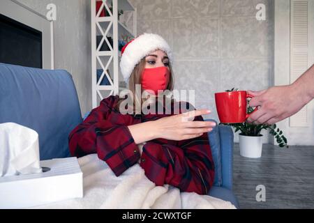 Gesundheit, Familie, Beziehung und Pflege Konzept. Mann, der der kranken jungen Frau rote Tasse mit heißem Tee gab, die auf dem Sofa in einer weißen Decke lag Stockfoto