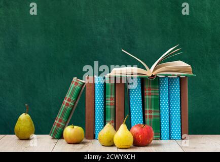 Offenes Lehrbuch, Stapel von Büchern in bunten Abdeckungen, Birnen und Äpfel auf Holztisch mit grünem Tafelhintergrund. Fernunterricht. Zurück zu s Stockfoto