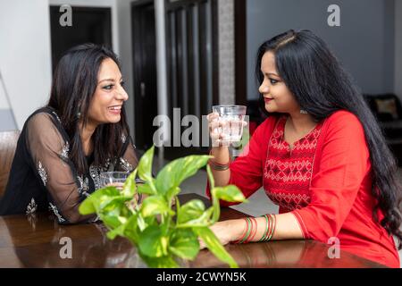 Portrait von zwei Freundinnen, die zu Hause sitzen und ein freundliches Gespräch führen, lächelnde Frauen, ein Glas Wasser halten und sich unterhalten. Stockfoto