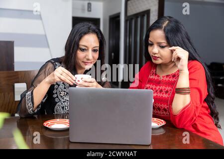 Zwei Freundinnen sitzen zu Hause mit Laptop vor ihnen, halten Teetassen und sprechen miteinander. Stockfoto