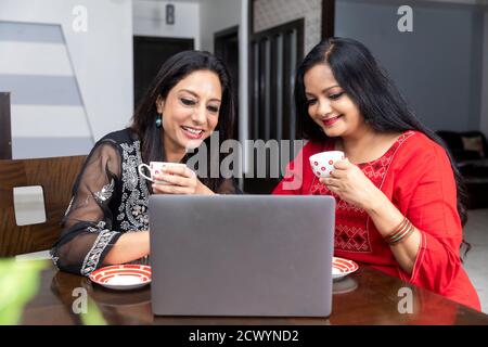 Zwei Freundinnen sitzen zu Hause mit Laptop vor ihnen, halten Teetassen und sprechen miteinander. Stockfoto