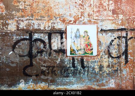 Varanasi, Uttar Pradesh, Indien : Nahaufnahme einer verwitterten Wand mit Devanagari-Schrift und einer Fliese, die die Götter Shiva, Parvati und Ganesh darstellt. Stockfoto