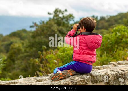 Shenandoah Valley, VA, USA 09/27/2020: Ein kleines kaukasisches Kind mit Mantel und Laufhose sitzt auf einer Steinwand von Skyline Drive Observin Stockfoto