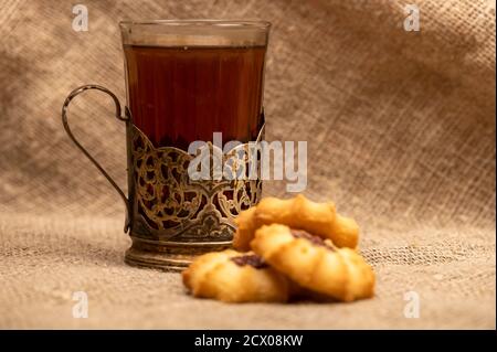 Hausgemachte Gebäck-Kekse mit Marmelade und einem facettierten Glas Tee in einem Vintage-Cup-Halter auf einem Hintergrund von homespun Stoff mit einer rauen Textur, close-up, Stockfoto