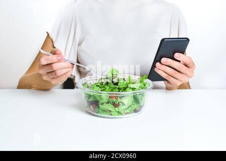 Nicht erkennbare kaukasische junge Frau, die Smartphone benutzt, während sie einen Salat aus einer Lunchbox isst. Konzept der Abhängigkeit von neuen Technologien Stockfoto