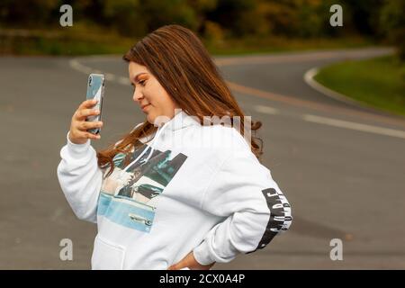 Shenandoah Valley, VA, USA 09/27/2020: Eine junge hispanische Frau mit braunem Haar im Kapuzenpullover macht ein Foto mit einem Smartphone. Sie hat ein Lächeln auf h Stockfoto