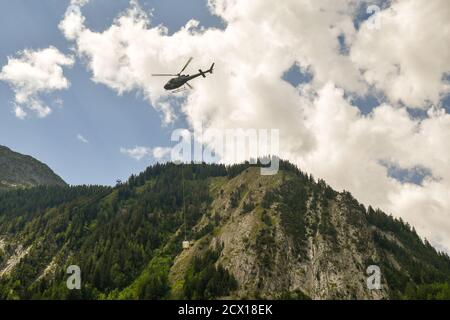 Helikopter mit Last auf der italienischen Seite des Mont Blanc Massivs gegen bewölkten Himmel im Sommer, Courmayeur, Aostatal, Italien Stockfoto