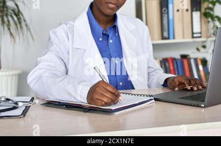 Afrikanische Frau hausarzt trägt weißen Mantel mit Laptop schreibt Notizen sitzt am Schreibtisch. Stockfoto