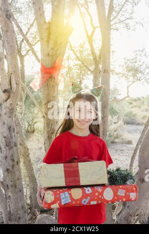 Ein Mädchen in rot gekleidet mit einem weihnachtlichen Stirnband, hält mehrere Kisten Weihnachtsgeschenke in ihren Armen, gegen das Licht des Sonnenuntergangs Stockfoto