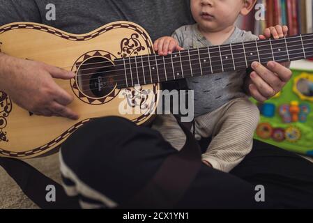 Papa auf dem Boden spielt kindergroße Gitarre, während er 1 Jahre alt auf der Runde hält. Stockfoto