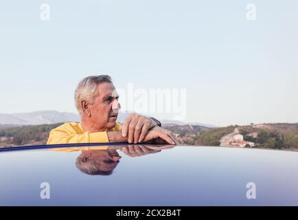 Älterer Mann, der sich auf das Fahrzeugdach lehnte und dabei wegschaute Roadtrip auf dem Land Stockfoto