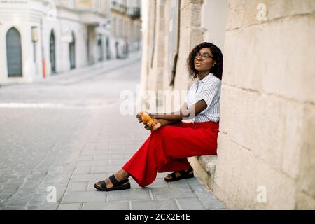 Horizontale Aufnahme von jungen attraktiven verträumt afrikanische Frau in roten Hosen und hellem Hemd gekleidet, sitzt auf der Treppe des alten Vintage-Gebäude auf der Stockfoto