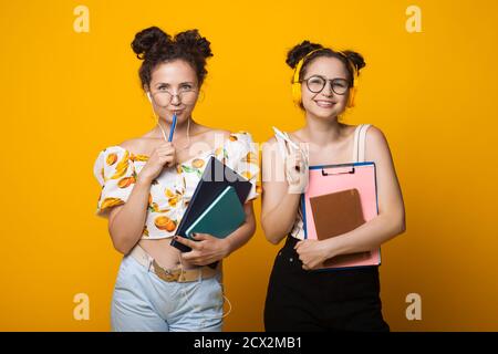 Die Zwillinge mit lockigem Haar lächeln durch eine Brille mit Ohrhörern auf die Kamera Und einige Ordner an einer gelben Wand halten Stockfoto