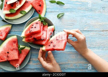 Draufsicht frische rote Wassermelone Scheibe in weiblichen Händen auf einem blauen rustikalen Holz Hintergrund. In Scheiben geschnittene reife Wassermelone mit Minze auf einem farbigen blauen Teller Stockfoto