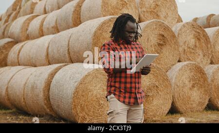 Junger afrikanischer Mann, der vor dem Heurollstapel an einer Tablette arbeitet. Smart Farming-Konzept. Hochwertige Fotos Stockfoto