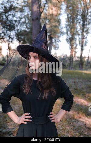 Hübsche junge schöne Frau in dunklem Kleid und Hexenhut in der Mitte des Herbstwaldes oder Park stehen. Halloween Party Kostüm. Nahaufnahme Porträt Stockfoto