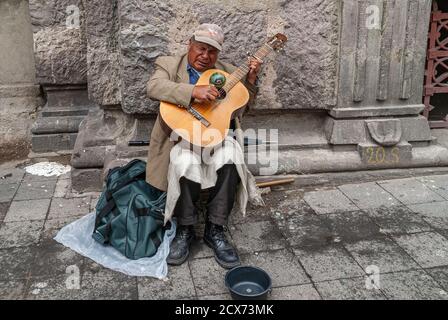 Quito, Ecuador - 2. Dezember 2008: Historische Innenstadt. Nahaufnahme eines älteren männlichen Gitarristen, der auf der Straße gegen die graue Steinfassade sitzt und um Mo bettelt Stockfoto