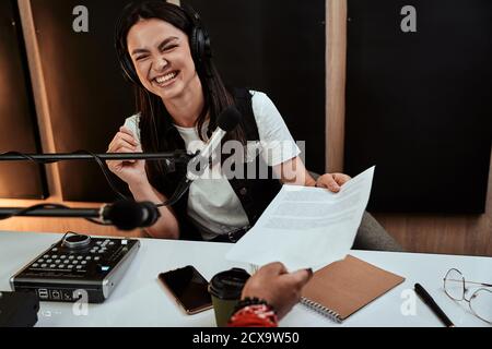 Portait von glücklicher Radiomoderatorin lächelt und erhält ein Drehbuch von ihrem männlichen Kollegen, während er eine Live-Show im Studio moderiert Stockfoto