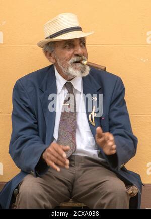 Trinidad, Kuba, 26. November 2017 - Der alte Mann sitzt auf der Bank das Rauchen einer Zigarre, gegen eine gelbe Wand Stockfoto