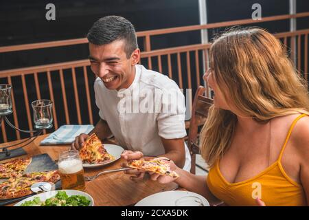 Gemischtes Rennpaar, das Pizza isst und laut lacht. Freunde essen und trinken zusammen Stockfoto