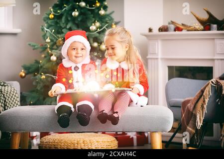 Süße kleine Kinder in Weihnachtsmannskostümen und mit Buch zu Hause am Vorabend Stockfoto