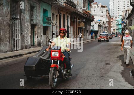 Straßenszene, Kubaner fährt ein traditionelles Motorrad mit Seitenwagen, gefolgt von einem Oldtimer. Mann in kubanischem T-Shirt auf der Seite (Havanna Kuba) Stockfoto