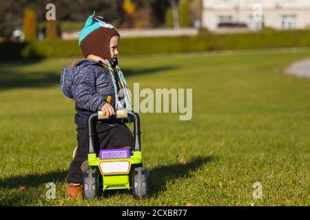 Kleiner Kleinkind Junge in einer dunklen Jacke und Hose spielt Im Park mit einem grünen Jeep Rollstuhl auf grün Gras in einem Stadtpark an einem warmen Herbsttag Stockfoto