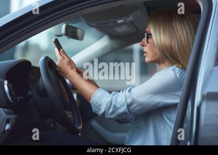Seitenansicht einer konzentrierten Frau oder Geschäftsdame Brillen sitzen hinter Lenkrad in ihrem modernen Auto und Mit dem Smartphone Stockfoto