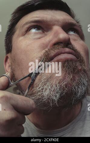 Ein erwachsener Mann schneidet sich mit einer kleinen Maniküre-Schere den Bart. Ungewöhnliches Porträt eines bärtigen Mannes mit grauem Haar. Grimassen vor der Kamera Stockfoto