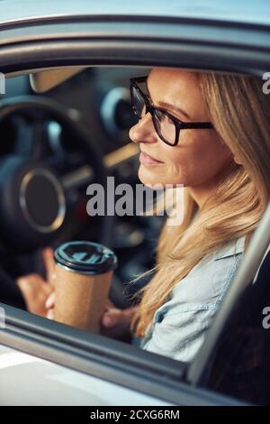 Kaffee zum trinken. Vertikale Aufnahme einer kaukasischen Geschäftsfrau mit Brillen, die hinter dem Lenkrad ihres modernen Autos sitzt und Kaffee hält Stockfoto
