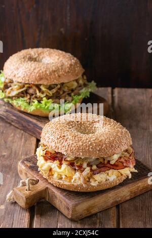 Zwei Bagels mit Rindereintopf, frischem Salat, Speck, Rührei und gebratener Zwiebel auf kleinen Holzbrettern auf einem Holztisch. Stockfoto