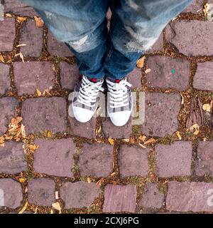 Die Beine eines jungen Mannes in Jeans und Tennis auf einem braunen kubischen Stein Hintergrund gekleidet. Fotos mit geringer Tiefe und strukturiertem Hintergrund. Stockfoto