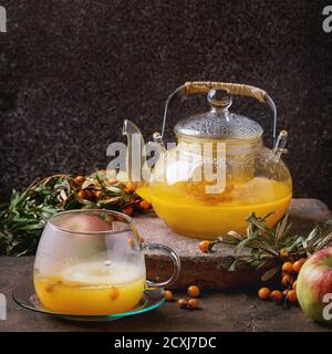Heißer Sanddorn-Tee-Getränk mit Äpfeln in Glas Teekanne auf Tontafel, serviert mit Glastasse, Untertasse, frische Beeren auf Zweig über alten hölzernen BA Stockfoto