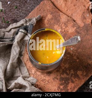 Dose Karottensuppe mit Löffel, stehend auf Küchenhandtuch und Tontafel über altem rostigen Eisen Hintergrund. Dunkler rustikaler Stil. Overhead-Ansicht. Quadratisch Stockfoto