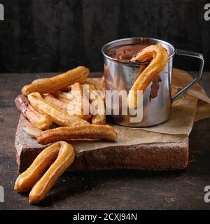 Traditionelle spanische Leckerbissen Churros mit Schokoladensauce in Metallbecher, serviert auf Backpapier auf dunklem Holzhintergrund. Quadratisches Bild Stockfoto