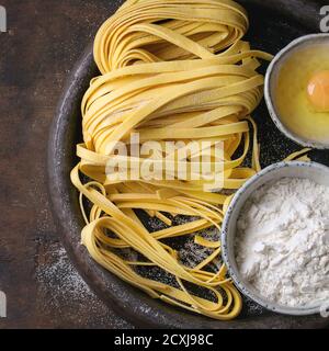 Rohe hausgemachte italienische Pasta-Tagliatelle mit Nudelschneider, Schüsseln mit weißem Mehl und gebrochenem Ei in alten Tonteller auf dunklem Holzboden. Stockfoto