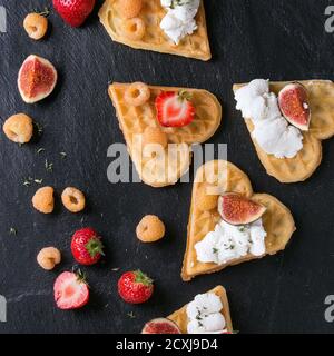 Waffeln als Herzform mit gelben Himbeeren, Erdbeeren, in Scheiben geschnittenen Feigen und Ricotta-Käse auf schwarzem Stein Schiefer Hintergrund. Draufsicht. Quadratisches Bild Stockfoto