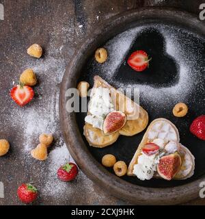 Waffeln als Herzform mit gelben Himbeeren, Erdbeeren, Feigen in Scheiben, Ricotta-Käse und Zuckerpulver auf Tonblech auf dunklem Holzhintergrund. Bis Stockfoto