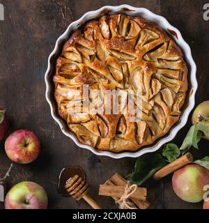 Hausgemachter Apfelkuchen in weißer Keramikform mit frischen Äpfeln mit Blättern, Honig und Zimtstangen auf dunklem Holzhintergrund. Draufsicht mit SPAC Stockfoto