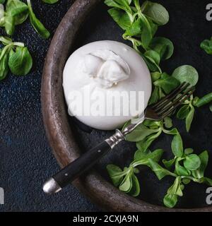 Ganze italienische Käseburrata mit Vintage-Gabel und frischem Mais-Salat in Tonblech über dunklem Strukturhintergrund. Overhead-Ansicht. Quadratisches Bild Stockfoto