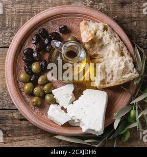 Grüne und schwarze Oliven mit Laib von frischem Brot, Feta-Käse, Flasche Olivenöl und junge Oliven Zweig auf Ton verzierten Teller über alten Holzbackgro Stockfoto