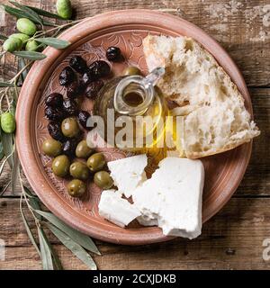 Grüne und schwarze Oliven mit Laib von frischem Brot, Feta-Käse, Flasche Olivenöl und junge Oliven Zweig auf Ton verzierten Teller über alten Holzbackgro Stockfoto