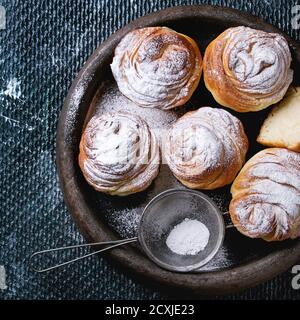 Moderne Backwaren Cruffins, ganze und Scheibe, wie Croissant und Muffin mit Zuckerpulver, serviert in Tonblech mit Vintage-Sieb über dunkle Textur backg Stockfoto