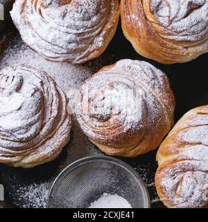 Moderne Backwaren-Cruffins, wie Croissant und Muffin mit Zuckerpulver, serviert in Tonblech mit Vintage-Sieb auf dunklem Texturhintergrund. Draufsicht, Stockfoto