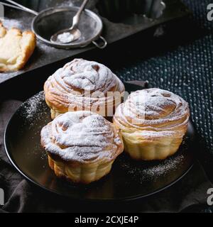 Moderne Backwaren Cruffins, ganz und in Scheiben, wie Croissant und Muffin mit Zuckerpulver, serviert auf schwarzem Teller mit Sieb, Silikonbackform und Text Stockfoto