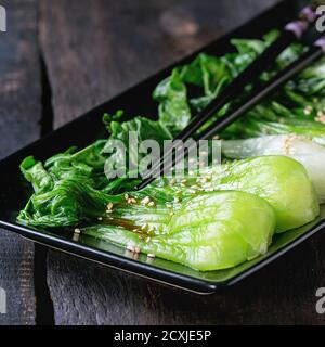 Gekochte bok Choy mit Sesam und Chili Pfeffer Olivenöl in schwarzem quadratischen Keramikplatte mit schwarzen Essstäbchen über alten Holztisch. Rustikaler Stil. Stockfoto